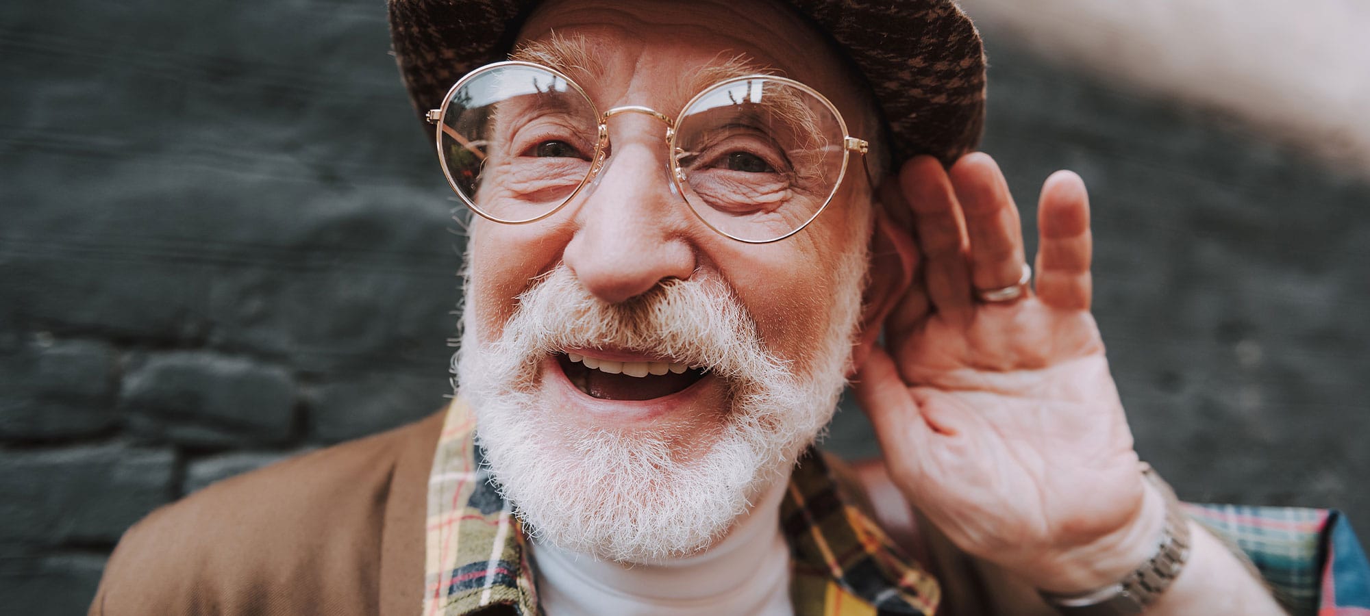 Älterer Herr, der lachend in die Kamera schaut und seine linke Hand ans Ohr hält, um zu symbolisieren, dass er zuhört.