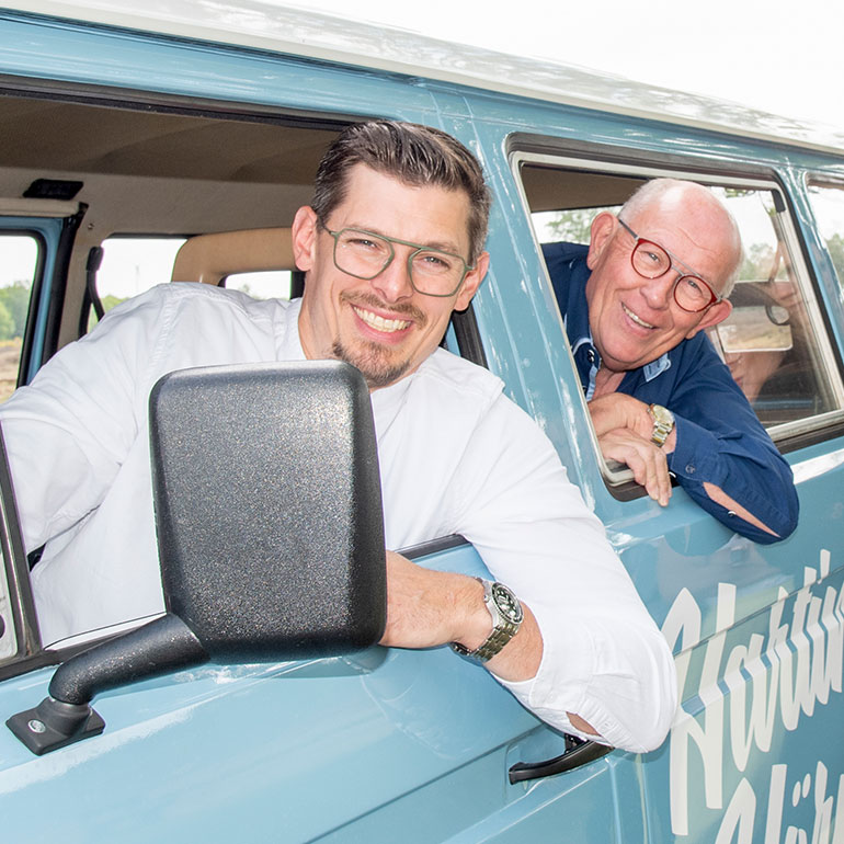 Christian und Eduard Harting schauen in der Halbtotalen aus dem Fenster eines blauen Vans – Sie trägen beide eine Brille.