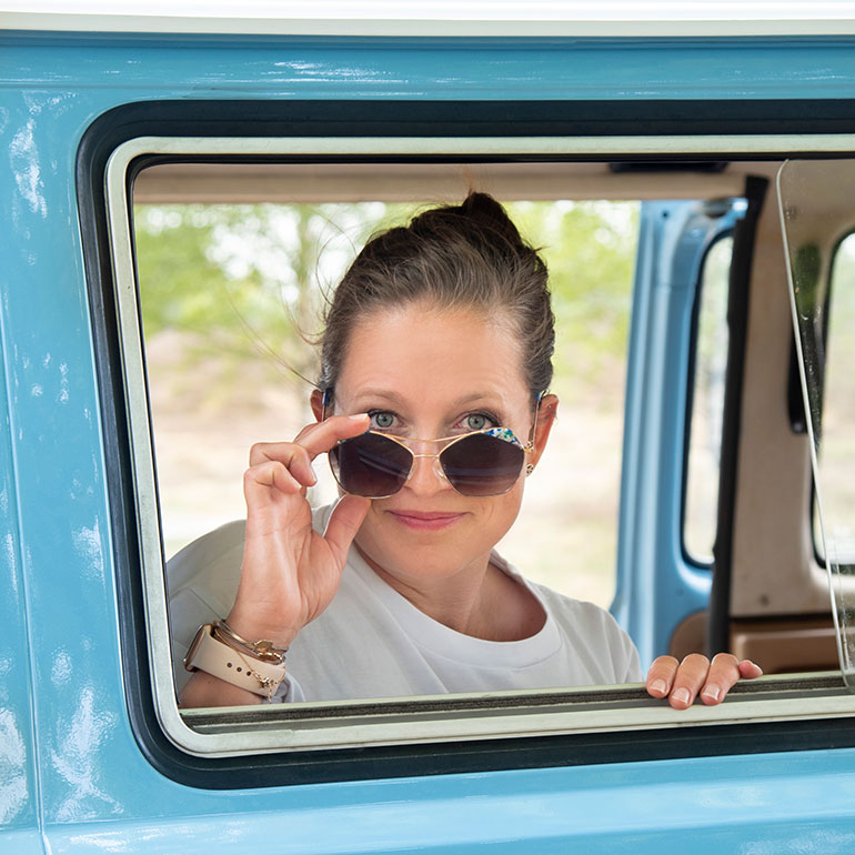 Nina Harting schaut in der Halbtotalen aus dem Fenster eines blauen Vans – Sie trägt eine Sonnenbrille und hält die das Gestell der Brille die Sie trägt in der rechten Hand.